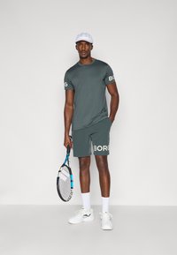 Athletic outfit featuring a dark green T-shirt and matching shorts with "BORG" branding. Includes a white cap and holding a tennis racket.
