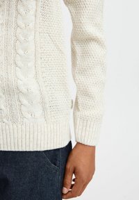 Suéter de punto grueso en crema con patrones de trenzado, puños y dobladillo acanalados. Combinado con denim oscuro, mostrando un diseño texturizado y cálido.