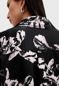 Camisa negra con estampado de grandes flores en blanco y gris; presenta un cuello, textura suave de satén y un detalle de botón en la parte posterior.