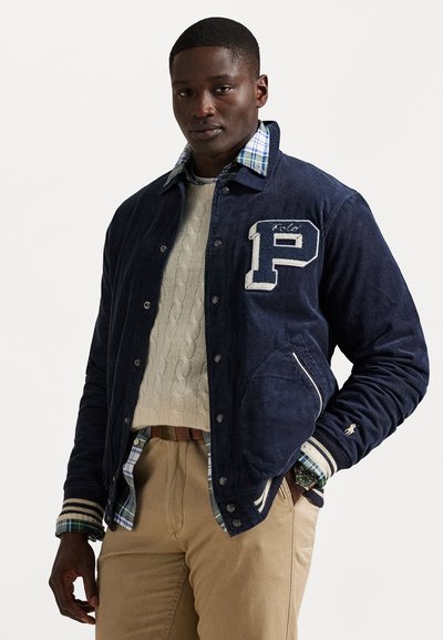 Navy corduroy jacket with a white "P" patch, striped cuffs, and a plaid-lined collar, layered over a cream cable-knit sweater and khaki pants.