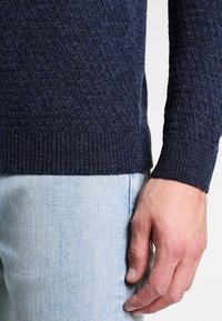 Homme portant un pull texturé bleu marine et un jean denim bleu clair, la main droite détendue le long du corps.