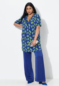 Blaue Tunika mit grünen und gelben kreisförmigen Mustern, kombiniert mit weiten blauen Hosen. Sie hat einen V-Ausschnitt und kurze Ärmel sowie einen silbernen Clutch.