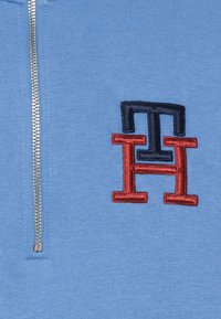 Ljusblå zip-up sweatshirt med en silverdragkedja och en broderad logotyp med röda och marinblå bokstäver "TH" och "H" i en staplad design.