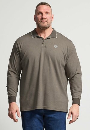 Mand med kort hår og skæg iført en brun langærmet polo og blå jeans, stående mod en ensfarvet lys baggrund.