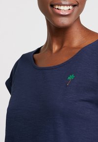 Camiseta de color azul marino en tejido suave con cuello redondo y mangas cortas. Presenta una pequeña palmera verde bordada en el pecho.