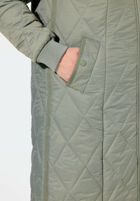 Gequilted lange jas in lichtgroen met ribbels aan de manchetten, voorzien van zijzakken en contrasterende afwerkingen langs de randen. Glanzende afwerking en ruitpatroon.