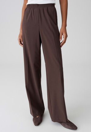 Frau trägt hochgeschnittene, weit geschnittene dunkelbraune Hose mit elastischem Bund und spitzen dunkelbraunen Schuhen, steht vor einem schlichten Hintergrund.