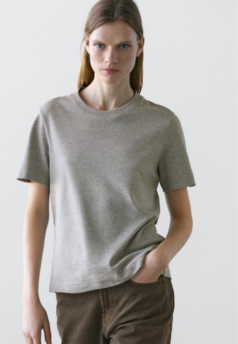 Jeune femme aux cheveux raides portant un t-shirt gris clair à manches courtes et un pantalon marron foncé, debout avec une main dans la poche.