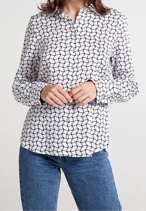 Donna con camicia bianca con bottoni e motivo circolare nero e jeans blu, mani intrecciate davanti in modo rilassato.