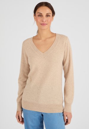 Maglione beige a collo a V realizzato in morbido tessuto a maglia, con maniche lunghe e orlo a coste. Indossato con pantaloni azzurri chiari.