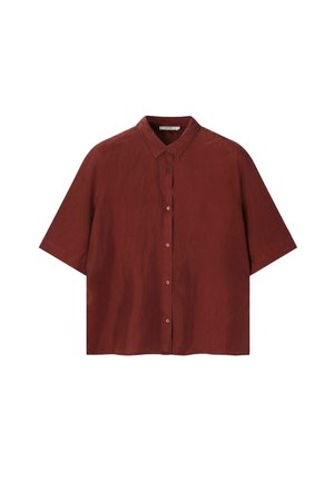 Blonde vrouw met schildpadzonnebril, losse roestkleurige blouse met knopen, brede lichtbruine ribbroek en bruine instapschoenen.