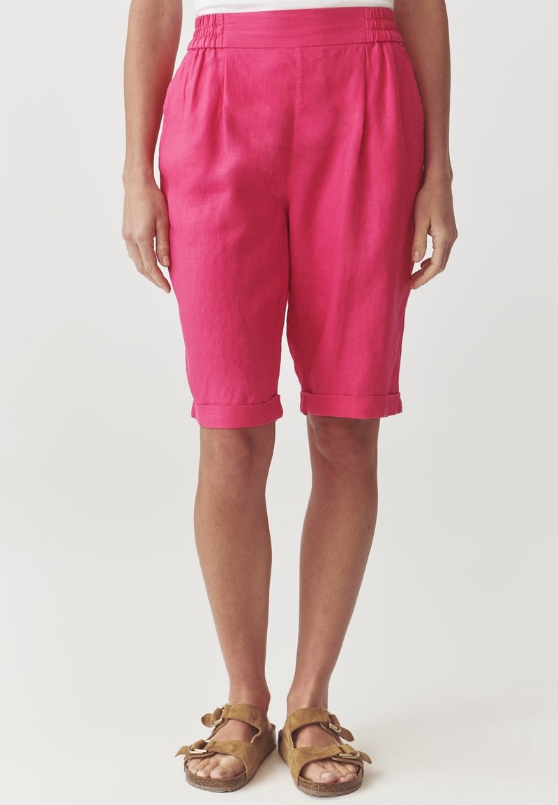 Femme portant un short rose vif jusqu'aux genoux avec des ourlets retroussés et des sandales marron à boucle et bouts ouverts, debout devant un fond blanc.