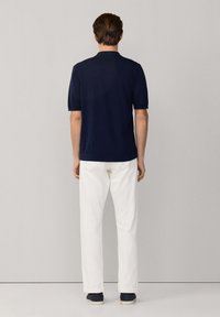 Hombre de espaldas, vestido con una camisa polo de manga corta azul marino, pantalones blancos y zapatos oscuros, de pie contra un fondo liso gris claro.