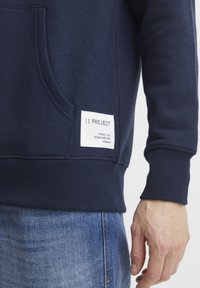 Navy blue sweatshirt made of soft fabric, featuring a front pocket, ribbed cuffs, and a white label with brand details on the lower side.