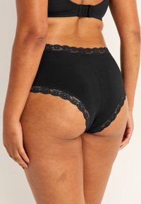 Högmidjade svarta underkläder tillverkade av mjukt tyg, med en spetsdetalj längs midjebandet och benöppningarna för extra detaljer.