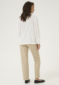 Top en dentelle blanche avec motif en losange, manches longues ; accompagné de pantalons beiges à coupe droite et de ballerines noires. Détail de tissu texturé visible.