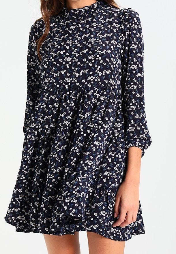 Marinblå blommig klänning med långa ärmar, volangkant och hög hals. Har mönster av vita och ljusblå blommor på lätt tyg.
