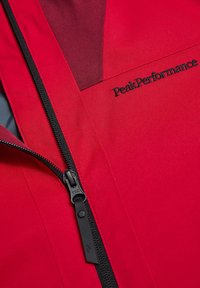Rød jakke med et glat, tekstureret stof, der har en sort lynlås og "PeakPerformance" logoet i sort på fronten.