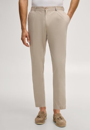 Mężczyzna w beżowych, dopasowanych spodniach i brązowych zamszowych mokasynach z chwostami, stojący z jedną ręką w kieszeni na jednolitym tle.