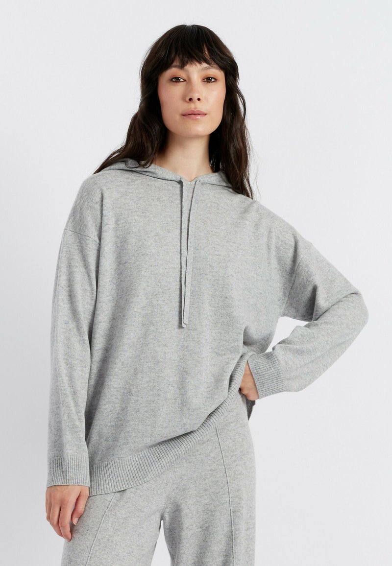 Grauer Kapuzenpulli aus weichem Strickstoff. Mit einem halben Reißverschluss am Hals, weiten Ärmeln und geripptem Saum. Kombiniert mit passenden Hosen.