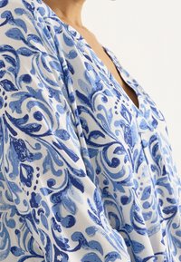 Blusa con estampado floral azul y blanco que presenta un escote en V profundo, mangas holgadas y una textura de tela suave y ligera.