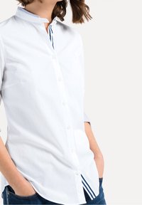 Bílá košile na knoflíky s vysokým límcem, se proužkovanými modrými akcenty na výstřihu a lemu, vyrobená z hladkého materiálu. Viditelné kapsy.