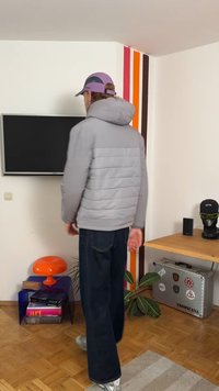 Szara kurtka puchowa z kapturem, pikowany wzór i zamek błyskawiczny z przodu. Połączona z ciemnymi dżinsami i trampkami, stojąc w pomieszczeniu z dekoracjami.