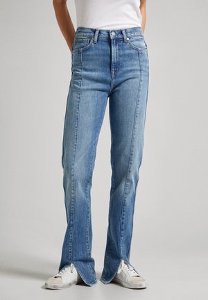 Persoon draagt lichtblauwe high-waist jeans met voornaad en gespleten zomen, gecombineerd met witte sneakers en een wit mouwloos topje.