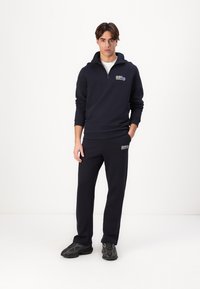 Felpa blu navy con mezza zip, polsini a costine e dettaglio del logo. Abbinata a pantaloni della tuta coordinati, tutto in tessuto morbido. Scarpe da ginnastica nere.
