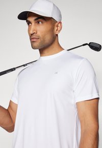 Bela športna majica s kratkimi rokavi, ki na prsih nosi subtilen logotip CK. Vidna je tudi svetla kapa in golf palica.