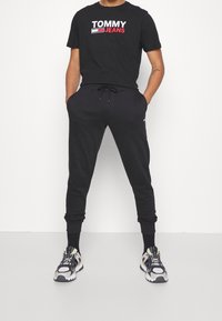 Černé bavlněné tričko s grafikou "TOMMY JEANS", spárované s černými tepláky se staženými manžetami a šnurkou. Sportovní tenisky dotvářejí vzhled.