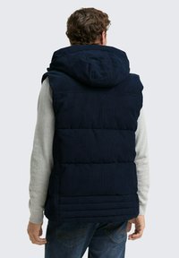 Vest cu umplutură de marina, cu textură ribbed, având o glugă și detalii de cusătură orizontală, purtat peste un tricou cu mânecă lungă de culoare gri.