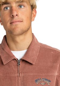Giacca in corduroy marrone con chiusura a zip, colletto a punta e logo ricamato sul petto. Tessuto testurizzato con righe verticali.