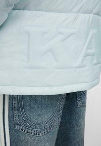 Šviesiai mėlynas pūkinis švarkas su iškeltu „KA“ logotipu, derinamas su šviesiu džinsiniu kelnių modeliu, turinčiu sudėtingą siuvinėjimą ir šonines juostas.