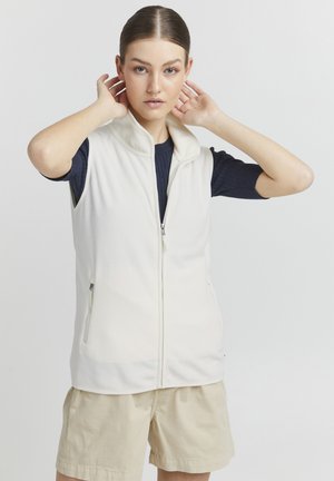 Cream fleece vest with a high collar, front zipper, and two side pockets, worn over a navy ribbed shirt and beige shorts.