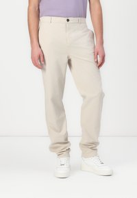 Pantalon beige à jambes droites avec une texture lisse, doté d'une fermeture à bouton et de poches latérales, associé à des baskets blanches.