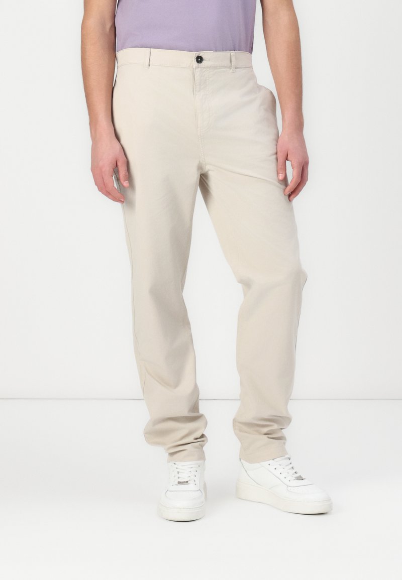 Pantalon beige à jambes droites avec une texture lisse, doté d'une fermeture à bouton et de poches latérales, associé à des baskets blanches.