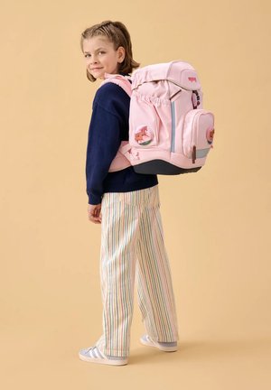 Junges Mädchen trägt gestreifte Hose und marineblauen Pullover und hat einen großen pinken Rucksack mit Pferde-Patches vor beigem Hintergrund.