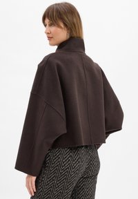 Bruine cropped jas met een hoge kraag en wijde mouwen, gemaakt van een gestructureerde stof. Een paar patroonrijke high-waisted broeken met een visgraatdesign.