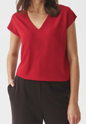 Kobieta w czerwonej bluzce z krótkim rękawem i dekoltem w serek oraz czarnych spodniach, stojąca z jedną ręką w kieszeni na jednolitym tle.