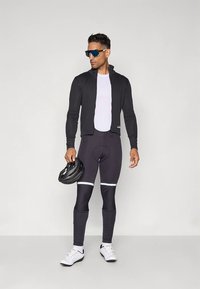 Άνδρας με μαύρο μπουφάν ποδηλασίας, μαύρο επενδυμένο κολάν με ανακλαστικές λωρίδες, λευκά παπούτσια, γυαλιά ηλίου, κρατώντας μαύρο κράνος, στέκεται σε λευκό φόντο.