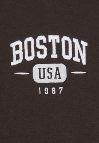 Tessuto marrone con testo ricamato bianco che dice "BOSTON USA 1987," con un font classico e pochi elementi decorativi.