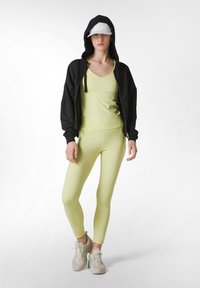Sort zip-up hættejakke, lysegrøn tætsiddende top og leggings, tekstureret stof, hvid kasket, beige sneakers med grønne accenter.