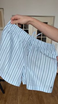 Világoskék rövidnadrág függőleges fekete csíkokkal és húzózsinóros derékkal, két kéz által tartva beltéren, háttérben fa padló.