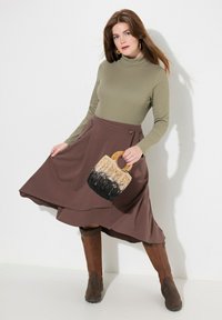 Bruine A-lijn rok met een taille knoop, gecombineerd met een aansluitende olijfgroene coltrui. Houdt een geweven tas met een lichte handgreep in handen.