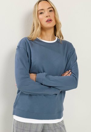 Blonde vrouw met gekruiste armen, gekleed in een blauw sweatshirt over een wit shirt en grijze geruite broek, staande tegen een lichte muur.