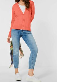 Korallfärgad cardigan med fem knappar, buren över en vit topp. Matchad med ljusblå jeans och vita sneakers, samt en flerfärgad halsduk.