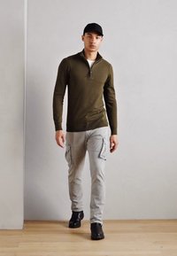 Haut zippé à manches longues olive, pantalon cargo gris clair avec poches, et bottes noires. Fond simple avec un sol en bois et des murs neutres.