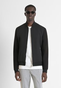 Giacca bomber nera con texture a costine sul davanti, maniche lisce, chiusura con zip e tasche laterali, abbinata a una maglietta bianca e pantaloni grigi.
