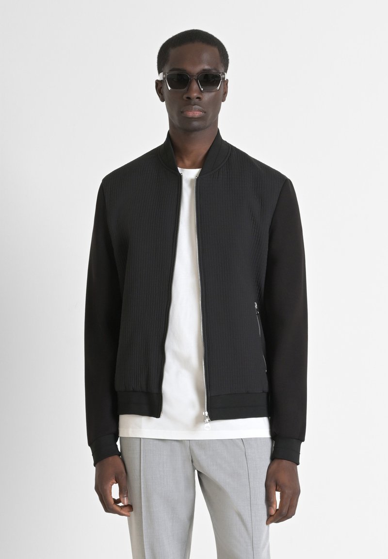 Giacca bomber nera con texture a costine sul davanti, maniche lisce, chiusura con zip e tasche laterali, abbinata a una maglietta bianca e pantaloni grigi.